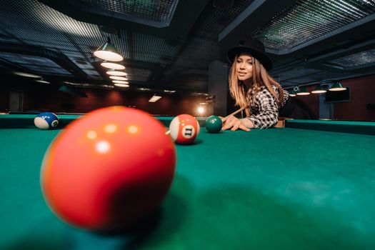 A girl in a hat in a billiard club with a cue in her hands hits a ball.Playing pool.