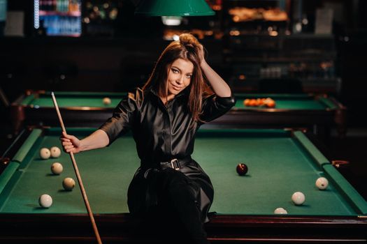 A girl with a cue in her hands sits on a table in a billiard club.Russian billiards.