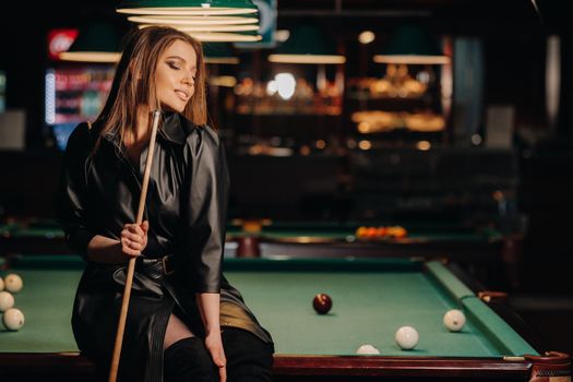 A girl with a cue in her hands sits on a table in a billiard club.Russian billiards.