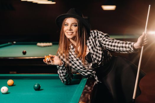 A girl in a hat in a billiard club with a cue and balls in her hands.Playing pool.