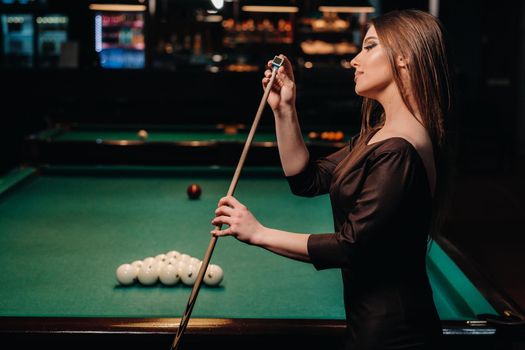 A girl in a dress stands with a cue in her hands and cleans it with chalk in a billiard club with balls in her hands.Playing billiards.