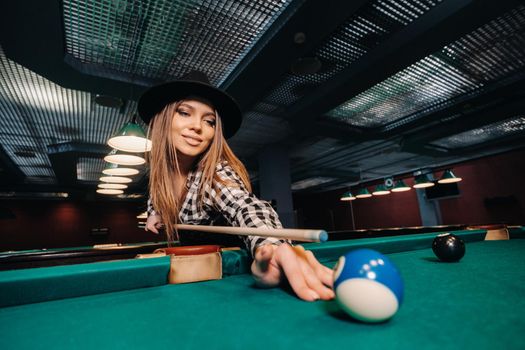 A girl in a hat in a billiard club with a cue in her hands hits a ball.Playing pool.