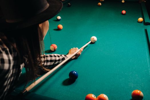 A girl in a hat in a billiard club with a cue in her hands hits a ball.Playing pool.