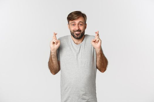 Portrait of hopeful smiling man with beard, wearing gray t-shirt, crossing fingers for good luck and looking at camera, making wish, standing over white background.