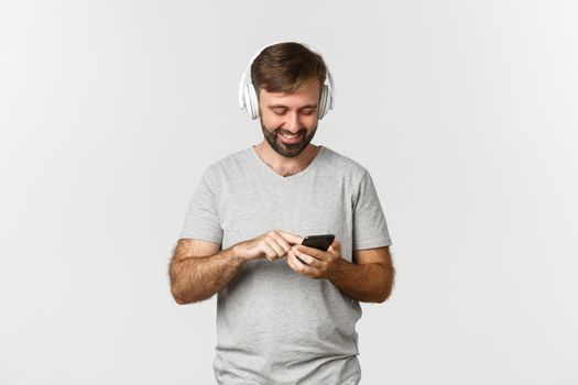 Portrait of attractive modern man smiling, looking at mobile phone, listening music in headphones, standing over white background.