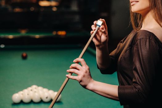 A girl in a dress stands with a cue in her hands and cleans it with chalk in a billiard club with balls in her hands.Playing billiards.