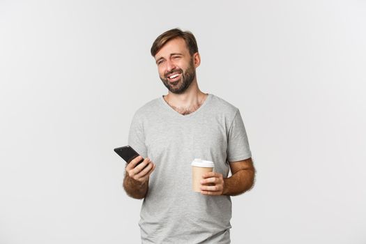 Image of modern caucasian man with beard, drinking coffee and casually using mobile phone, laughing as if talking to someone, standing over white background.