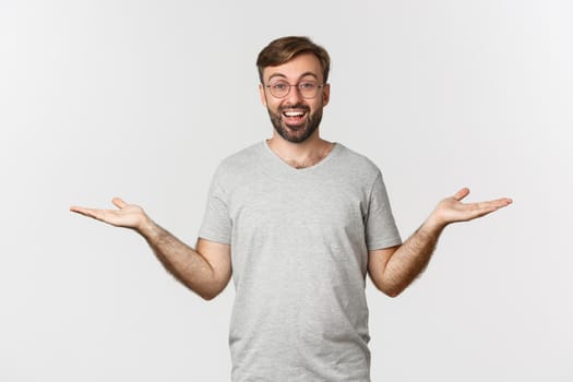 Handsome man with beard, wearing gray t-shirt and glasses, spread hands sideways and holding two things, demonstrate products, standing over white background.