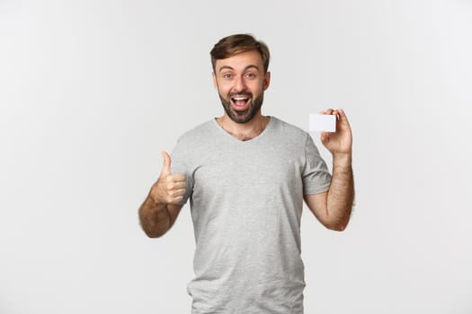 Portrait of handsome smiling man in gray t-shirt, showing credit card, making thumbs-up in approval, standing over white background.