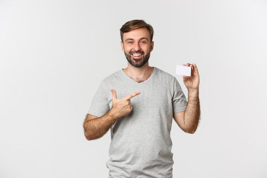 Portrait of handsome bearded man in gray t-shirt, pointing at credit card and smiling, standing over white background.