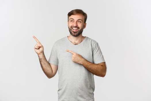 Handsome smiling man in gray t-shirt, looking dreamy at upper left corner, showing logo, standing over white background.
