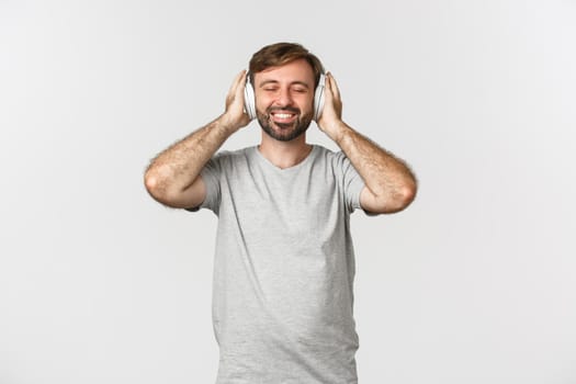 Image of handsome man in casual outfit, listening music in wireless headphones, smiling pleased, standing over white background.