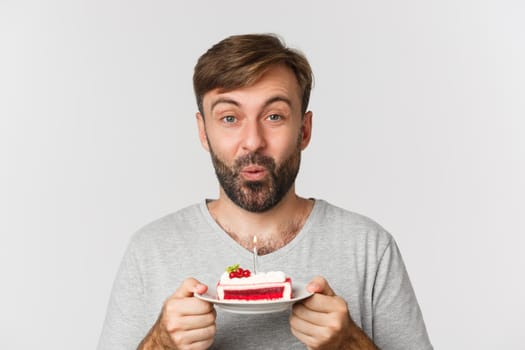 Close-up of excited bearded man, smiling and celebrating birthday, holding cake with lit candle, making b-day wish, standing over white background.