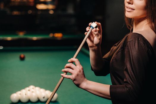 A girl in a dress stands with a cue in her hands and cleans it with chalk in a billiard club with balls in her hands.Playing billiards.