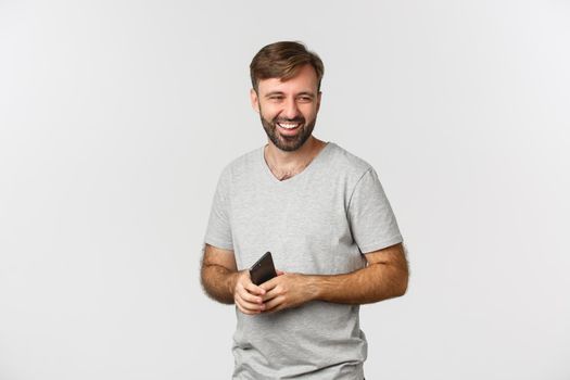 Image of handsome bearded man in gray t-shirt, holding smartphone, looking right and laughing, standing over white background.