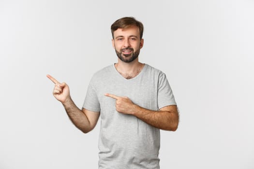 Handsome adult man with beard, wearing grey t-shirt, pointing fingers left and smiling, showing your logo, standing over white background.
