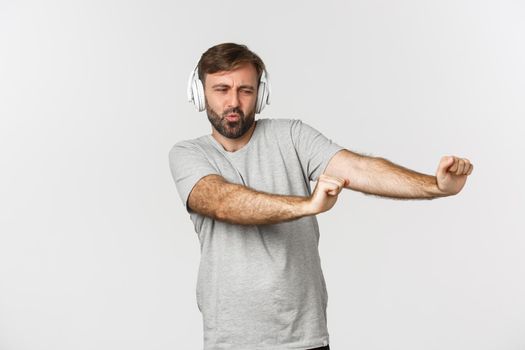 Portrait of happy guy dancing to music in headphones, standing over white background.