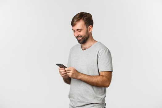Portrait of modern bearded man in gray t-shirt, messaging and looking at smartphone happy, standing over white background.