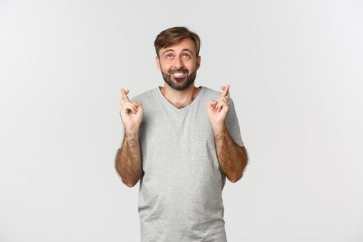 Hopeful smiling man in gray t-shirt, looking up and making wish, crossing fingers for good luck, standing over white background.