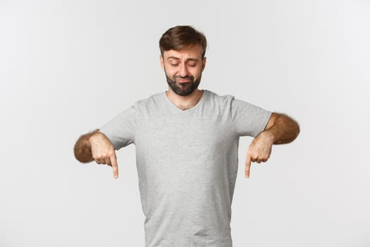 Portrait of skeptical bearded man in gray t-shirt, frowning and pointing fingers down, showing something bad, standing over white background.