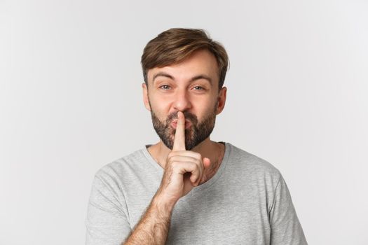 Close-up of smiling man in gray t-shirt, hushing, telling a secret, standing over white background.