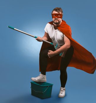 Superhero ready for cleaning work. Man in a mask and superhero red cloak posing with a bucket and a mop presenting himself as a rock musician. Isolated on a blue background.