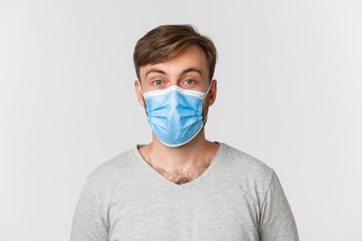 Concept of pandemic, covid-19 and social-distancing. Close-up of surprised man in medical mask, raising eyebrows amazed, standing over white background.