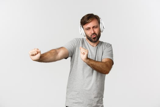 Portrait of happy handsome guy in gray t-shirt, listening music in wireless headphones and dancing, standing over white background.