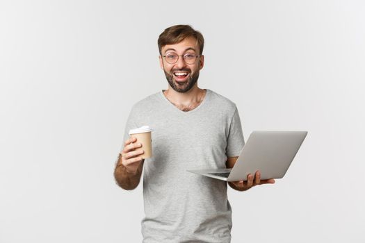 Portrait of handsome male freelancer, drinking coffee and working with laptop, standing over white background.