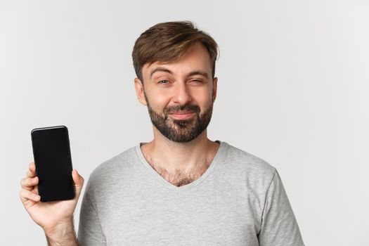 Close-up of pleased smiling man with beard, looking delighted and showing mobile phone screen, standing over white background.