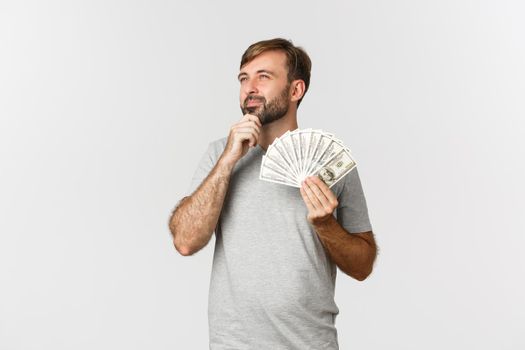 Thoughtful nice guy with beard, holding money and thinking about shopping, looking at upper left corner.