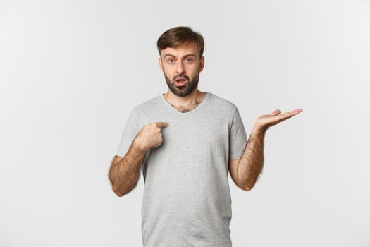 Portrait of handsome bearded man in gray t-shirt, pointing at himself and shrugging confused, being accused, standing over white background.