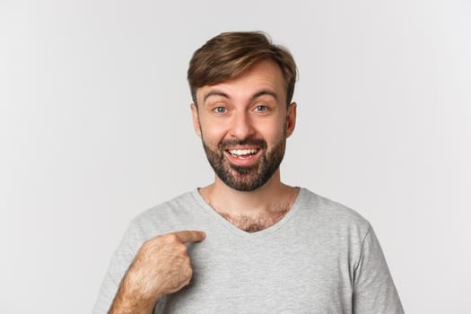 Close-up of surprised handsome man with beard, pointing at himself and smiling, standing over white background.