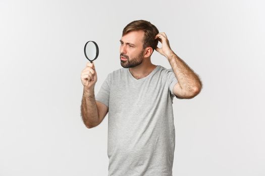 Image of handsome caucasian man in gray t-shirt, searching for something, looking confused through magnifying glass with curious face, standing over white background.