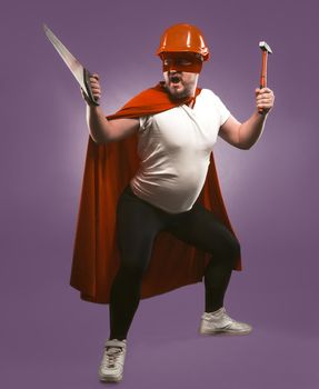 Super hero man holding construction tools. Engineer or Repairman in red helmet and super hero uniform with saw and hammer standing isolated on grape purple background.