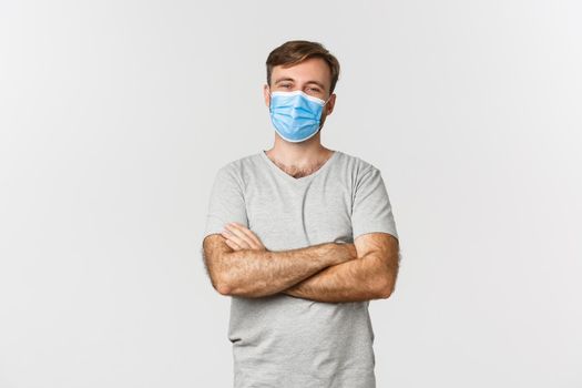 Concept of pandemic, covid-19 and social-distancing. Handsome and confident man in medical mask, cross arms on chest and smiling, standing over white background.