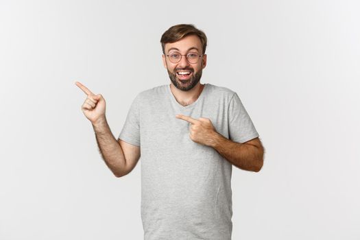 Excited bearded man smiling, pointing fingers at upper left corner, showing logo, standing over white background.