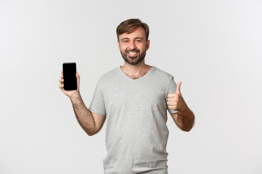 Image of handsome man in gray t-shirt, showing mobile phone screen and thumbs-up in approval, like something good, standing over white background.
