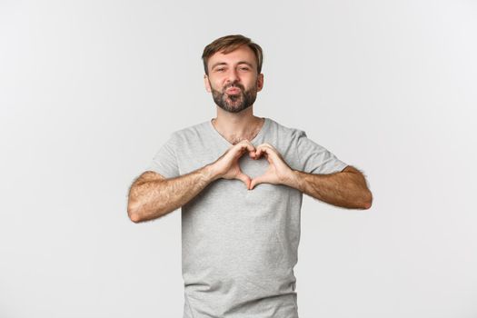 Portrait of handsome smiling man with beard, showing heart sign, confess in love, kissing someone, standing over white background.