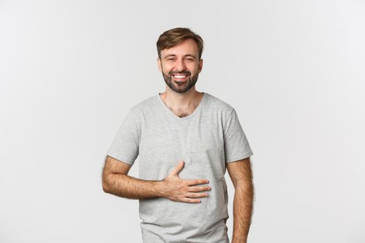 Portrait of happy man feeling pleased after eating tasty food, rubbing belly and smiling delighted, standing over white background.