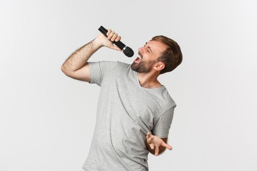 Handsome carefree guy singing song in karaoke, holding microphone, standing over white background.