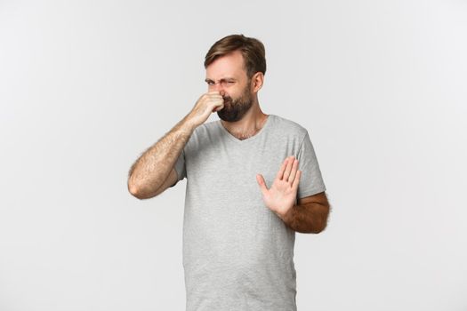 Image of an ordinary guy in gray t-shirt shut his nose, rejecting something disgusting with awful smell, standing over white background.