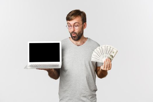 Handsome male freelancer showing laptop screen and money, standing over white background.