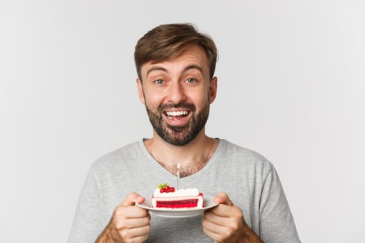 Close-up of excited bearded man, smiling and celebrating birthday, holding cake with lit candle, making b-day wish, standing over white background.