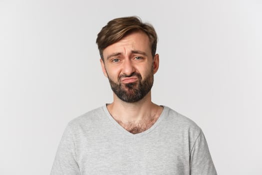 Close-up of gloomy disappointed guy with beard, wearing gray t-shirt, grimacing and frowning sad, standing over white background.