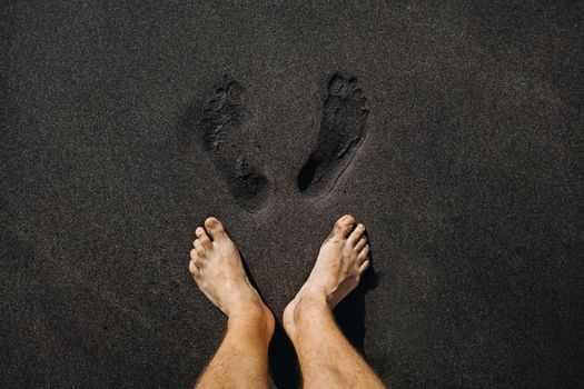 Close up of male footprints and feet walking on the volcanic black sand on the beach.