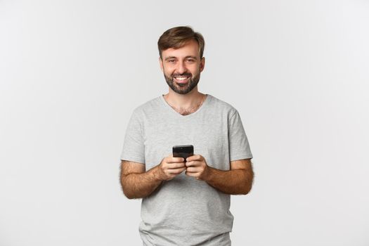 Portrait of happy smiling man with beard, wearing t-shirt, using mobile phone, standing over white background.