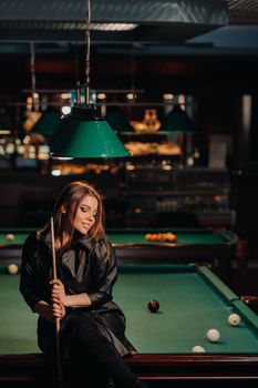 A girl with a cue in her hands sits on a table in a billiard club.Russian billiards.