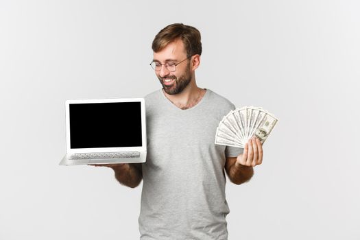 Handsome male freelancer showing laptop screen and money, standing over white background.