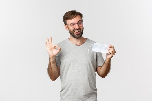 Concept of pandemic, coronavirus and social-distancing. Image of handsome smiling guy in glasses, showing okay sign and recommending to wear medical mask.